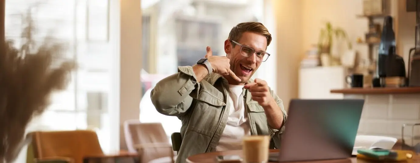 Retrato de un chico alegre y coqueto con gafas, sentado con una computadora portátil en una cafetería, indicando una llamada telefónica.